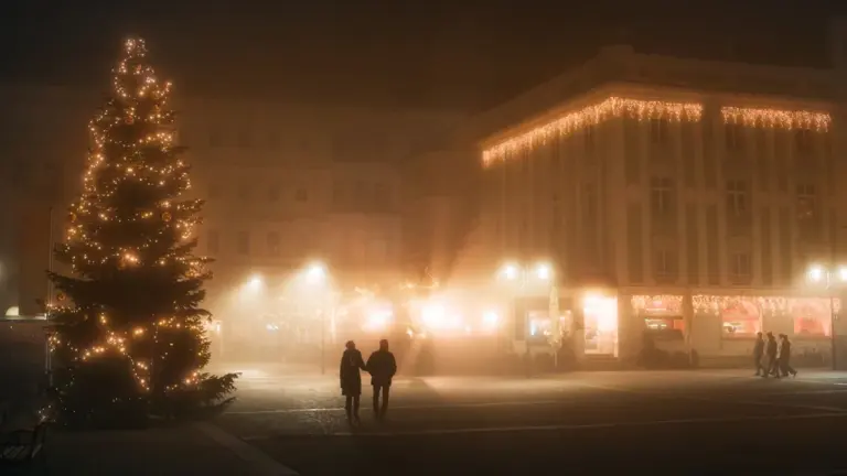 Ein beleuchteter Weihnachtsbaum auf einem nebligen Stadtplatz mit einem spazierenden Paar am Abend.