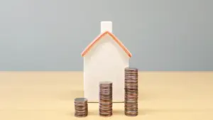Ein kleines Hausmodell neben aufgetürmten Münzen auf einem Holztisch als Finanzsymbol.