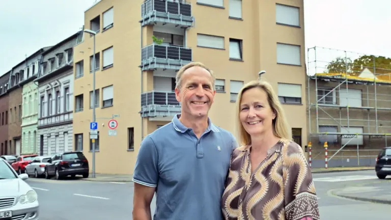 Ein lächelndes Ehepaar steht vor einem modernen beigen Wohnhaus an einer belebten Straße.