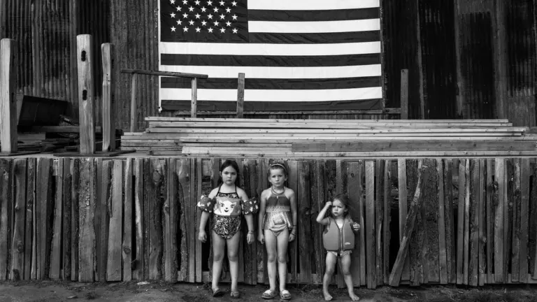 Drei kleine Mädchen stehen barfuß vor einer Holzwand mit einer hängenden amerikanischen Flagge.
