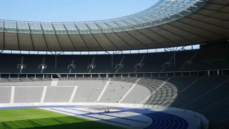Weitwinkelaufnahme des Berliner Olympiastadions mit blauer Laufbahn und grüner Rasenfläche bei Tageslicht.