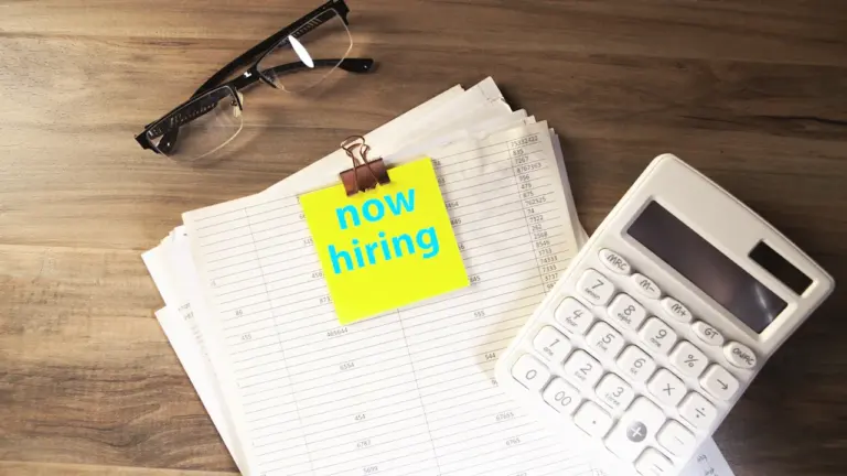 Ein Taschenrechner und Notizen auf einem Schreibtisch mit einer 'Now Hiring'-Notiz.