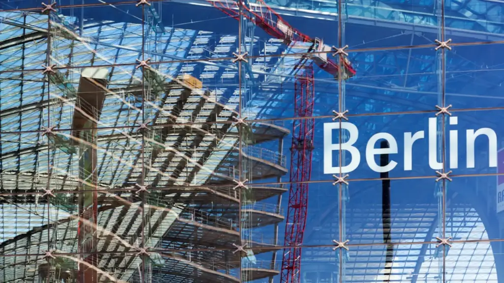 Moderne Glasfassade mit Berlin-Schriftzug und Baustellenkränen in einer Reflexion.